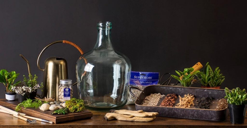 materials laid out for creating an indoor bottle garden, including glass bottle, soil, gravel, and small plants.